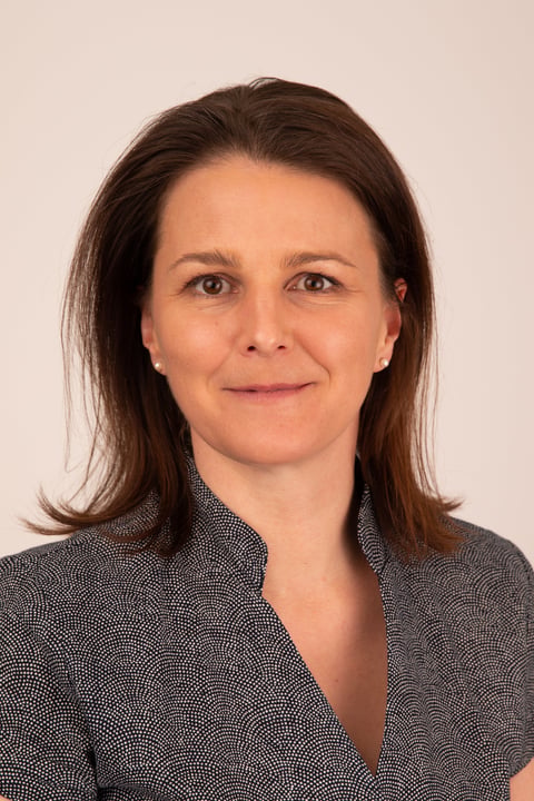 Professional headshot of a woman with long dark brown hair wearing a dark patterned blouse against a neutral beige background