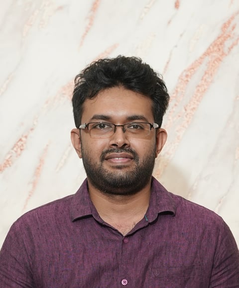 Man with black hair, glasses, and beard wearing a purple button-up shirt against a light marble background