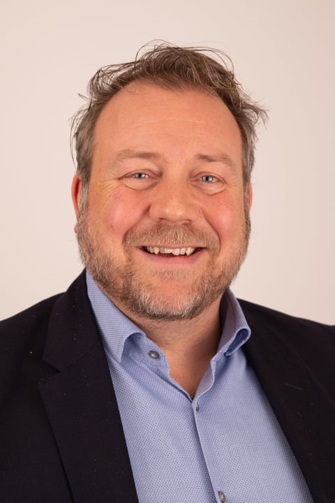 Headshot of a smiling middle-aged man with brown hair and beard wearing a dark blazer over a light blue shirt against a neutral background