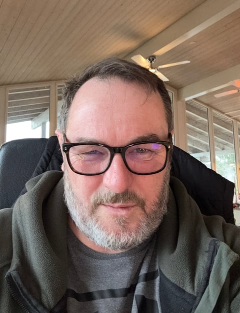 Middle-aged man with glasses and beard wearing a dark jacket, sitting indoors with wooden ceiling and ceiling fan visible